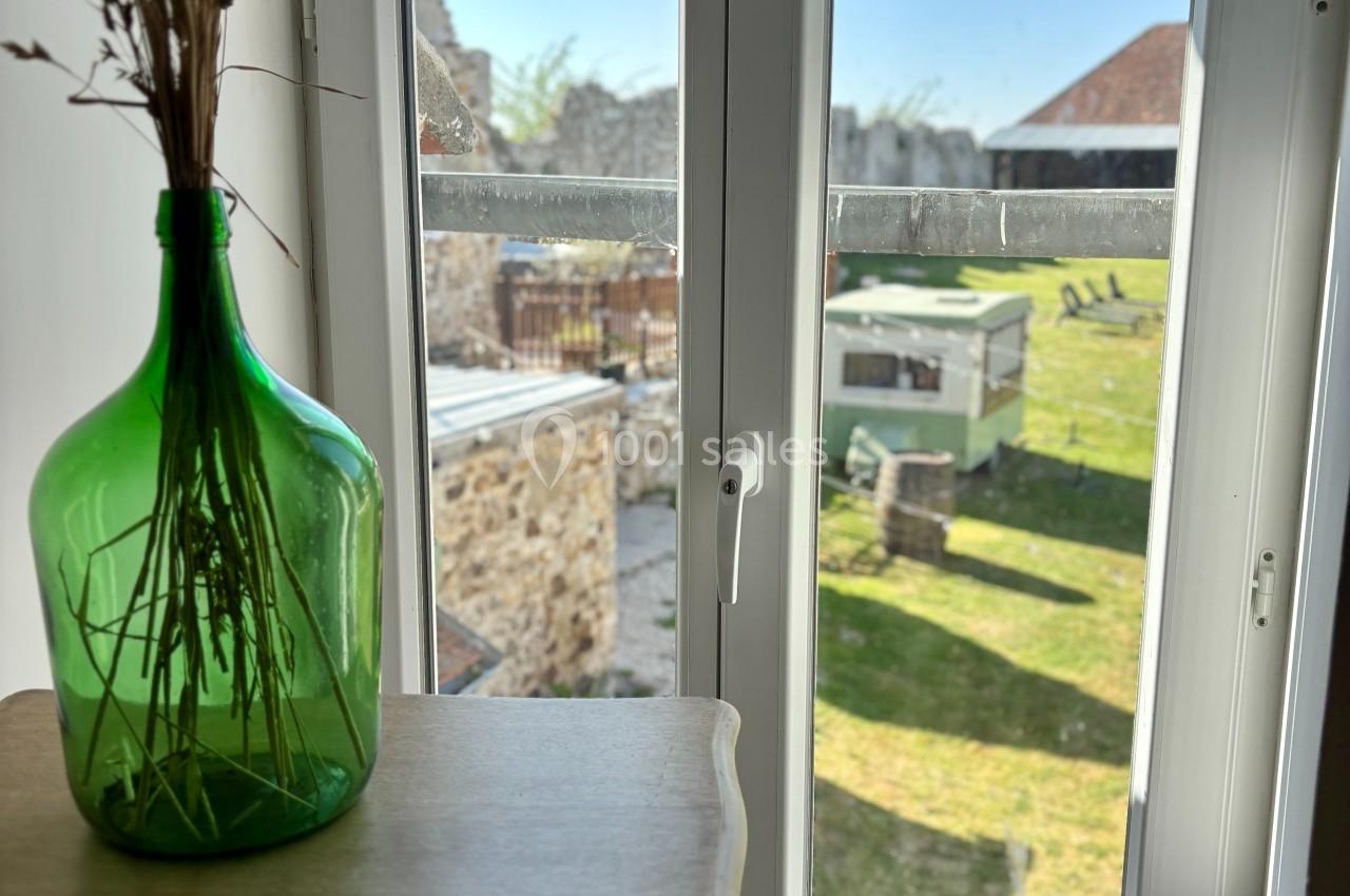 Vue depuis une fenêtre donnant sur un jardin ensoleillé avec une pelouse, des bâtiments et un vase vert contenant des herbes…