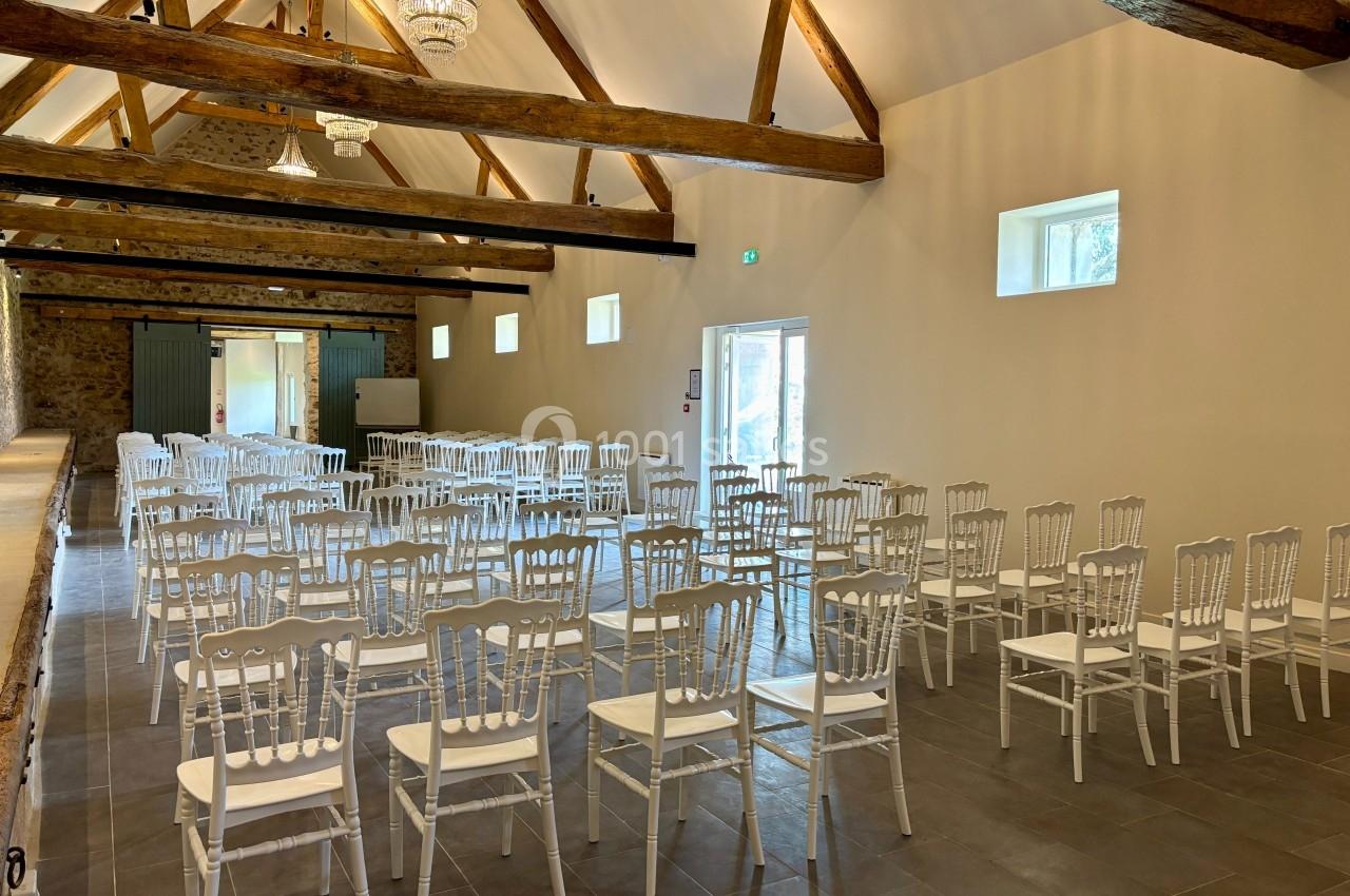 Salle lumineuse avec des chaises blanches alignées, poutres apparentes au plafond et murs en pierre.