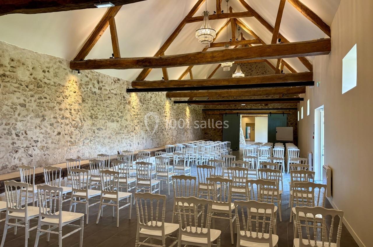 Salle avec poutres apparentes, murs en pierre et rangées de chaises blanches disposées pour un événement.