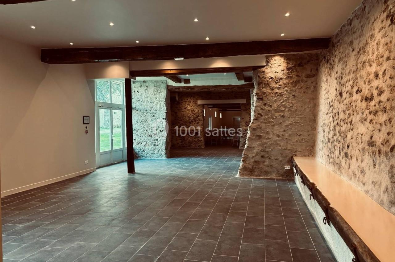 Salle intérieure lumineuse avec murs en pierre apparente, poutres en bois et sol en carrelage gris.