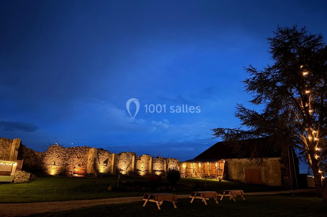 Ruines éclairées et bâtiment adjacent sous un ciel nocturne, avec un arbre illuminé et des bancs sur une pelouse.