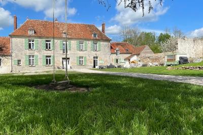 Miniature La cour du Domaine Ruines éclairées et bâtiment adjacent sous un ciel nocturne, avec un arbre illuminé et des bancs sur une pelouse.