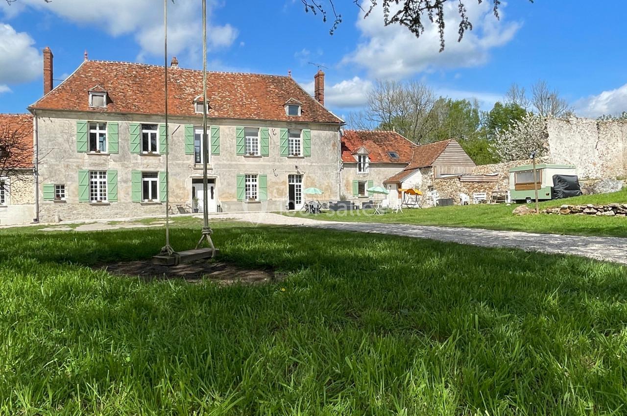 Grande maison ancienne en pierre avec toit en tuiles rouges, entourée d'un jardin verdoyant sous un ciel partiellement…