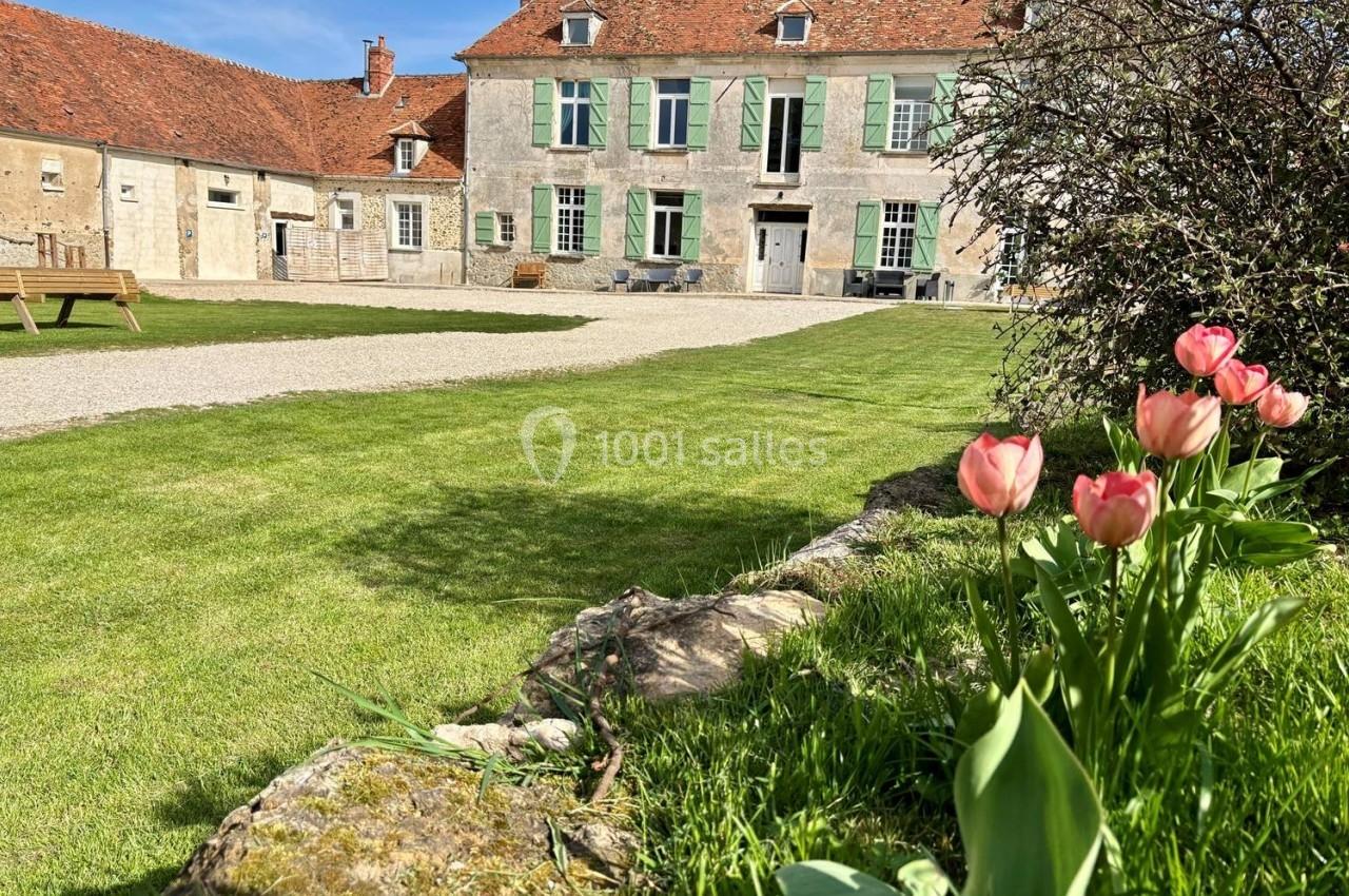 Jardin verdoyant avec des tulipes roses au premier plan et une maison ancienne en pierre avec volets verts en arrière-plan.