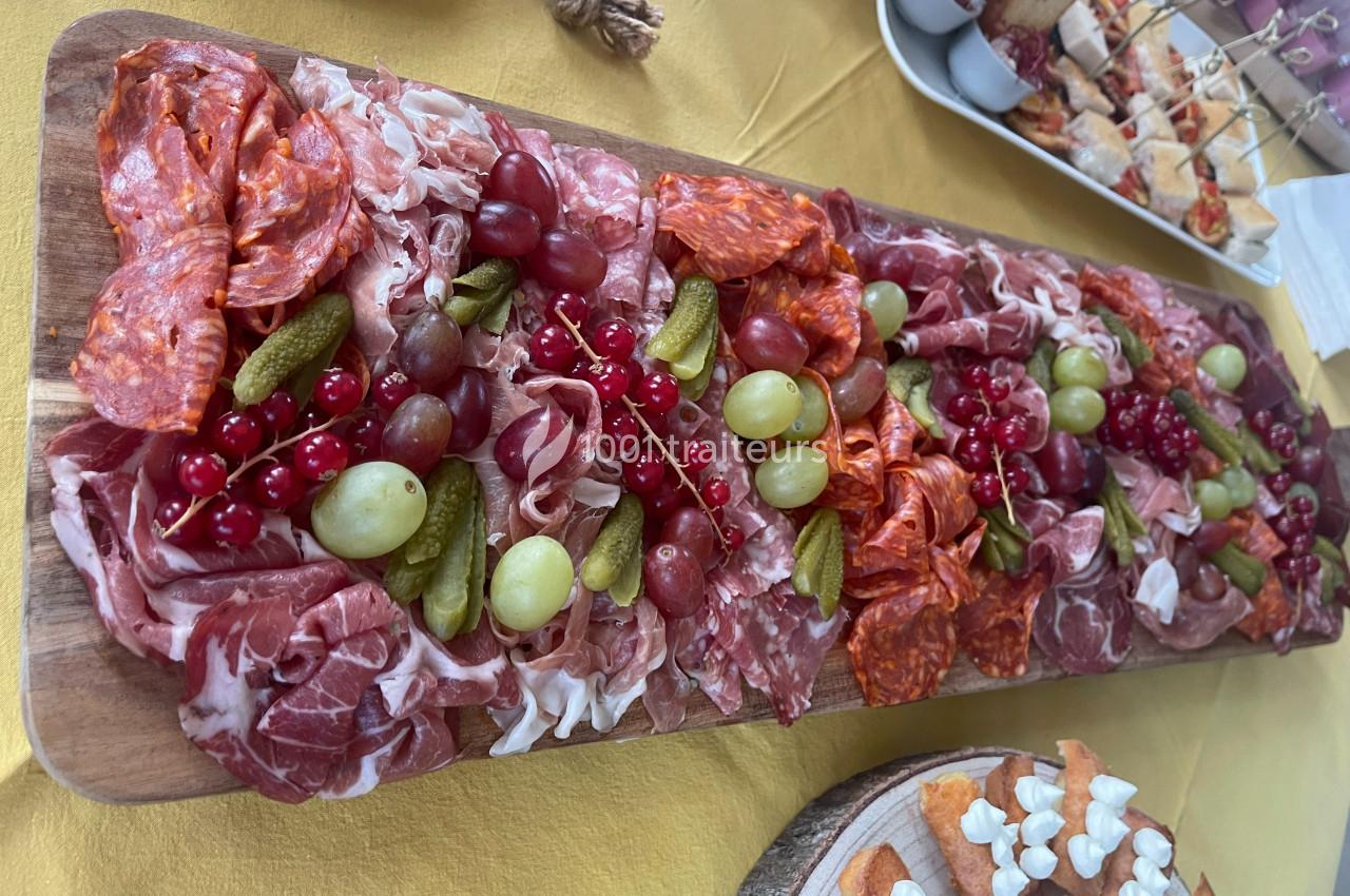 Plateau de charcuterie variée avec raisins, groseilles et cornichons, présenté sur une table jaune.