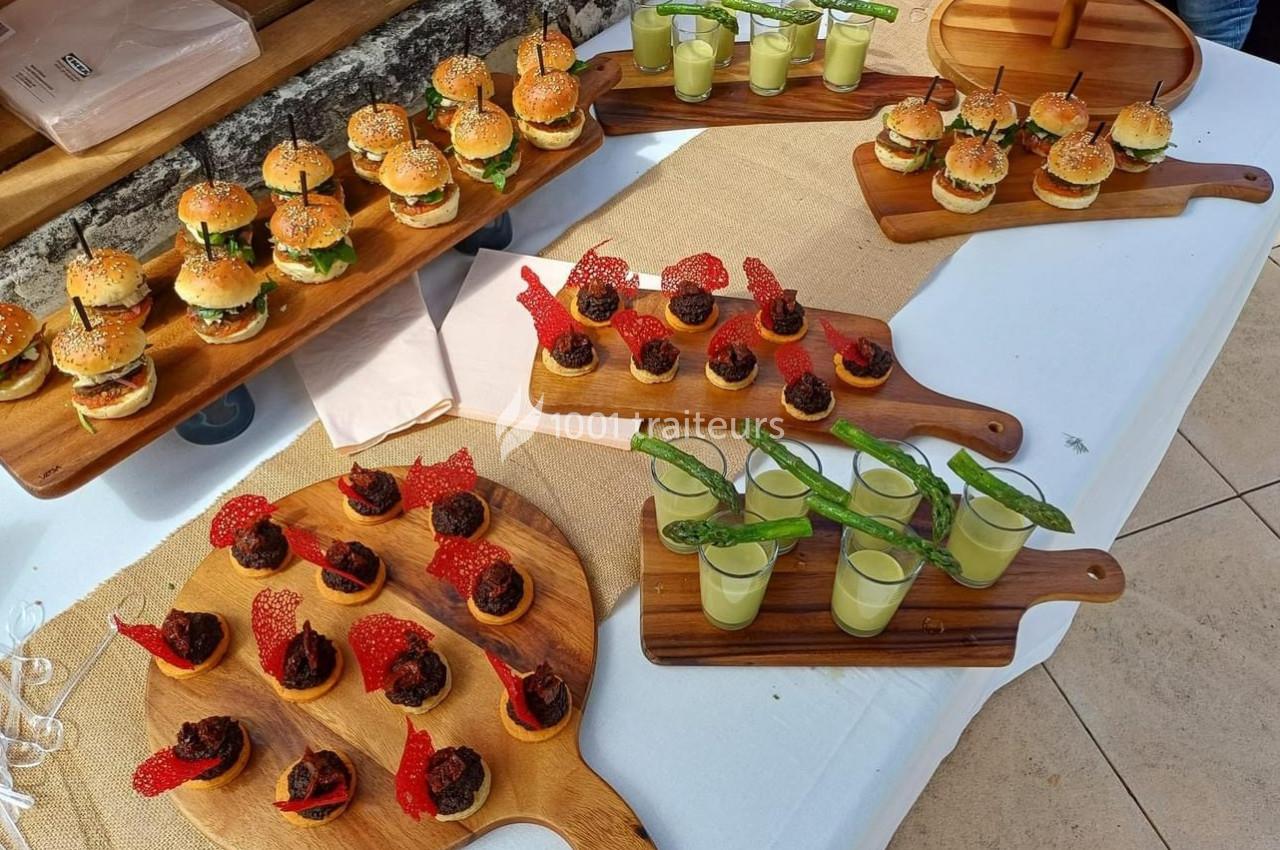Plateaux en bois garnis de mini-burgers, verrines vertes et bouchées décorées de tuiles rouges sur une table blanche.