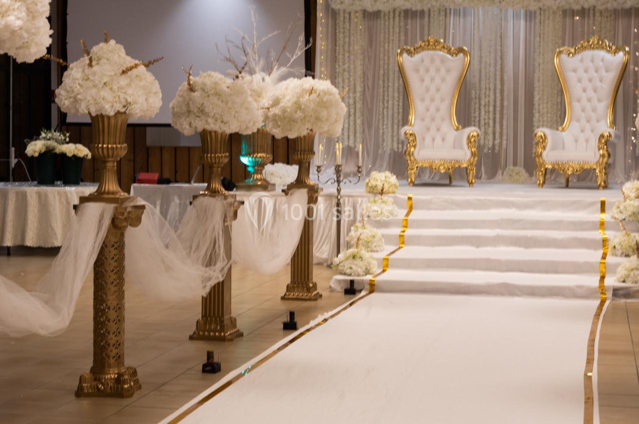 Allée décorée de colonnes dorées et de fleurs blanches menant à deux fauteuils royaux sur une scène.