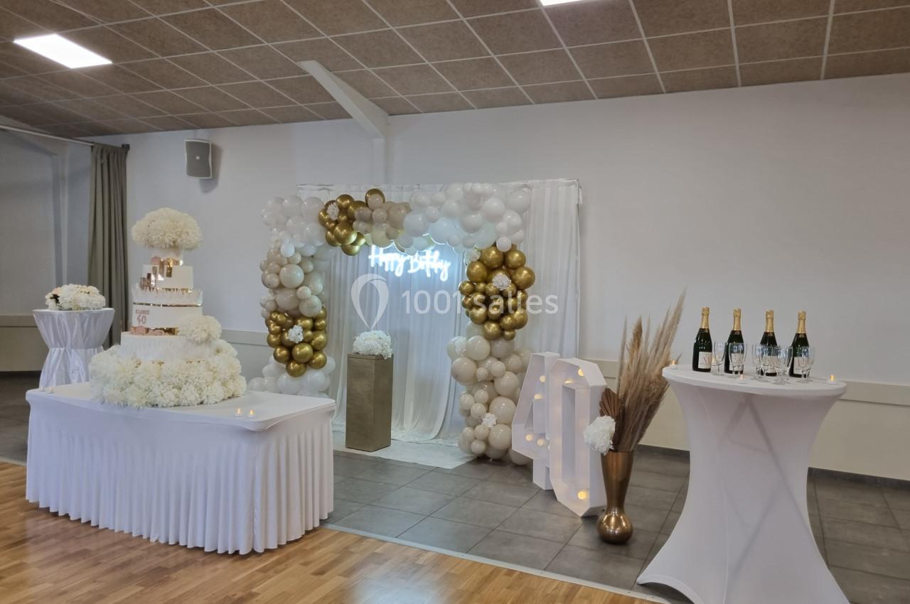 Décoration de fête avec arche de ballons dorés et blancs, table avec gâteau et bouteilles sur des tables hautes.