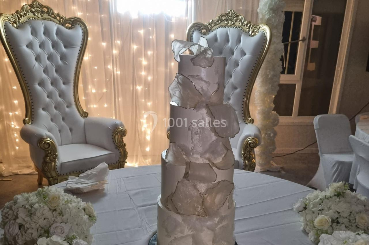 Gâteau de mariage à plusieurs étages décoré de pétales blancs, devant deux fauteuils ornés et un fond lumineux.