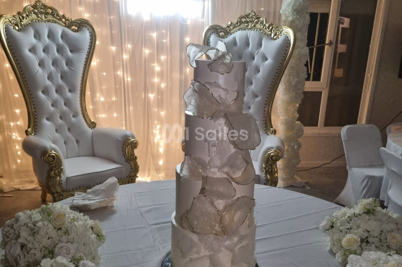 Gâteau de mariage à plusieurs étages décoré de pétales blancs, devant deux fauteuils ornés et un fond lumineux.