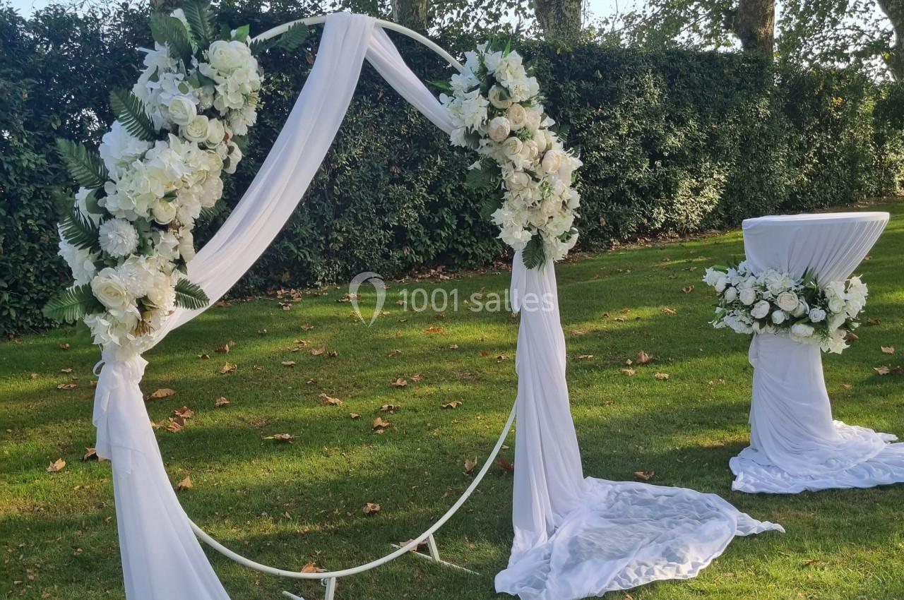 Arche circulaire décorée de fleurs blanches et de voilages, installée sur une pelouse avec une table assortie.