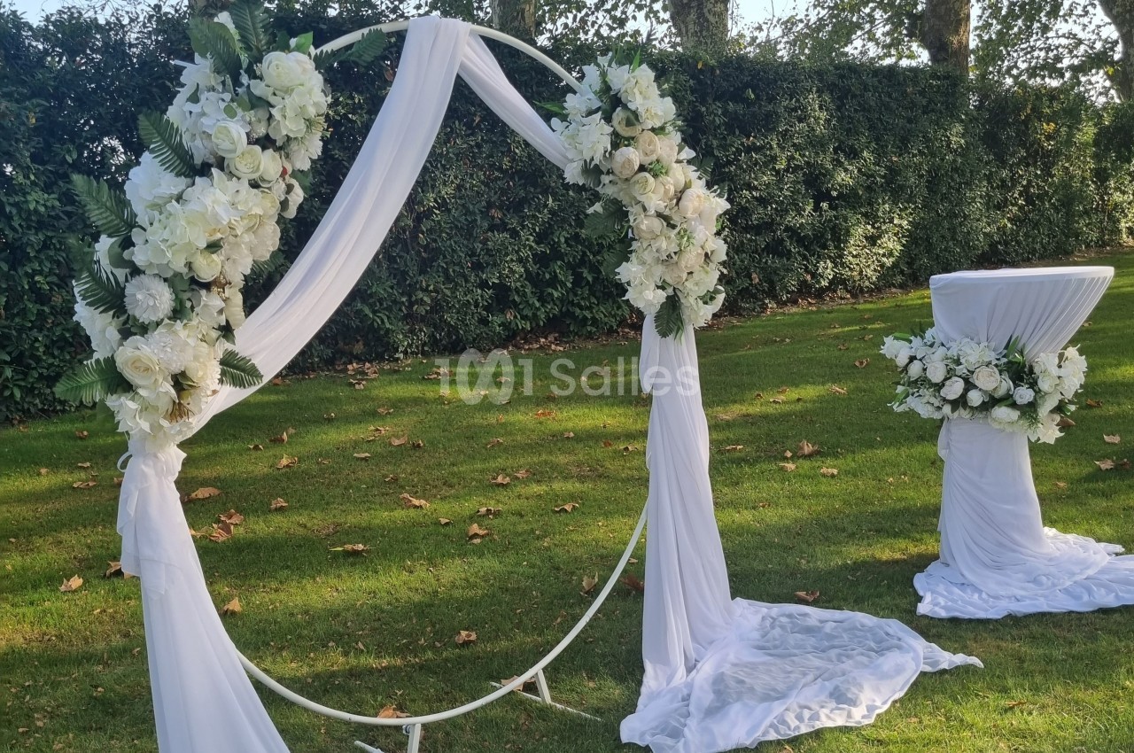 Arche circulaire décorée de fleurs blanches et de voilages, installée sur une pelouse avec une table assortie.