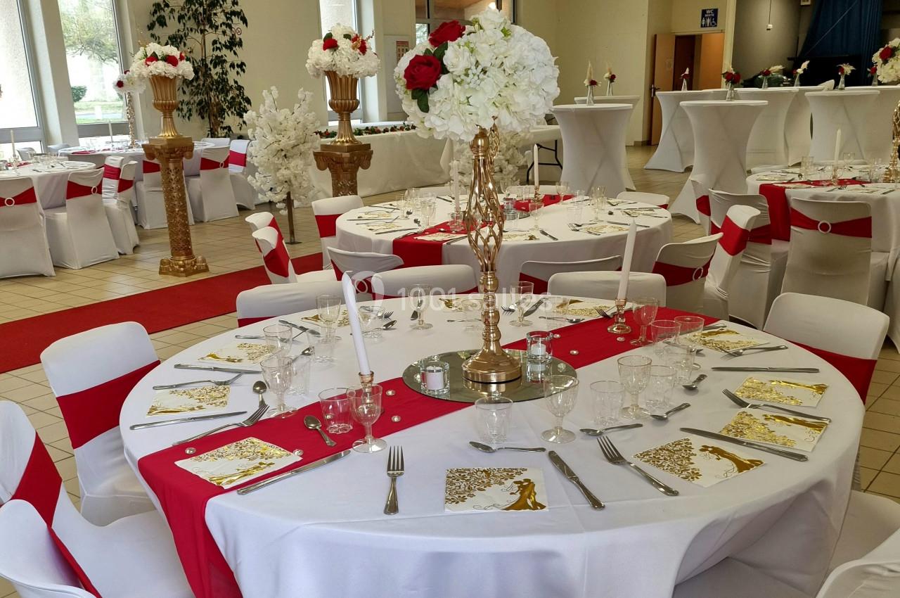 Salle décorée pour un événement avec tables rondes dressées, nappes blanches, chemins de table rouges et centres de table…