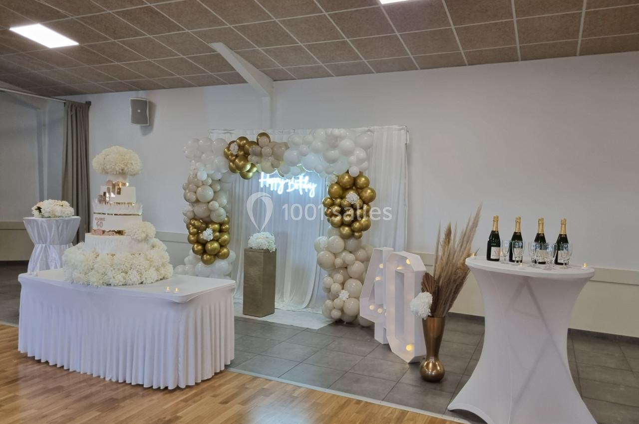 Décoration de salle de fête avec arche de ballons blancs et dorés, gâteau sur une table et bouteilles de champagne.