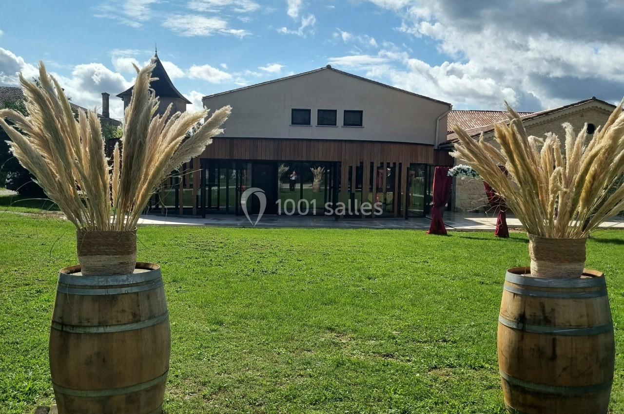 Deux tonneaux décorés de pampas encadrent une pelouse devant un bâtiment moderne sous un ciel partiellement nuageux.