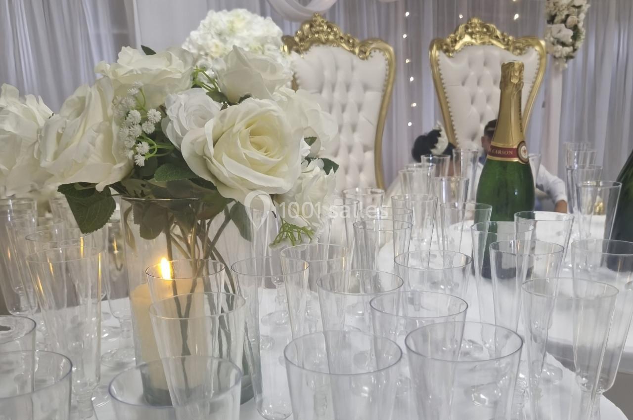 Arrangement de verres transparents, bouquet de fleurs blanches, chandelle et bouteille de champagne sur une table élégante.