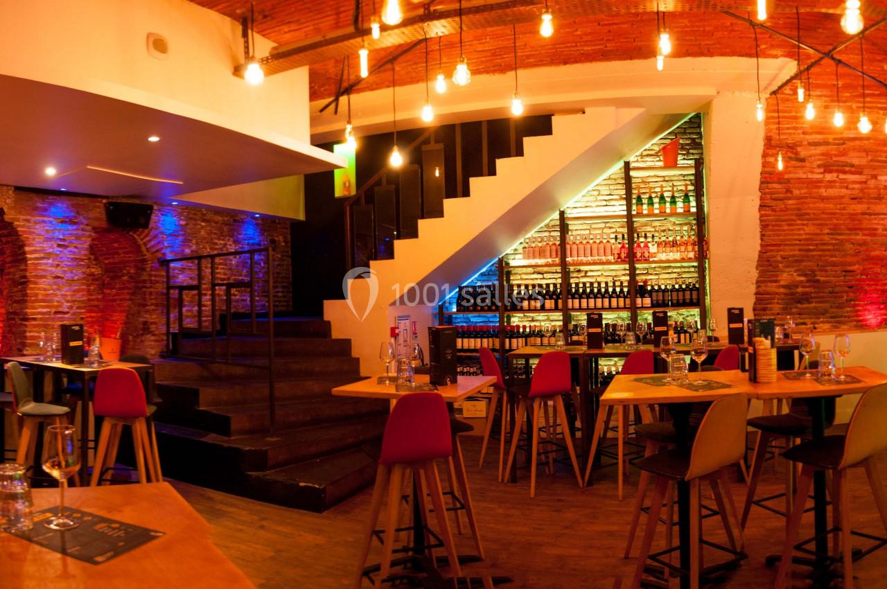Salle de bar avec tables hautes, chaises colorées, éclairage chaleureux et étagères remplies de bouteilles.