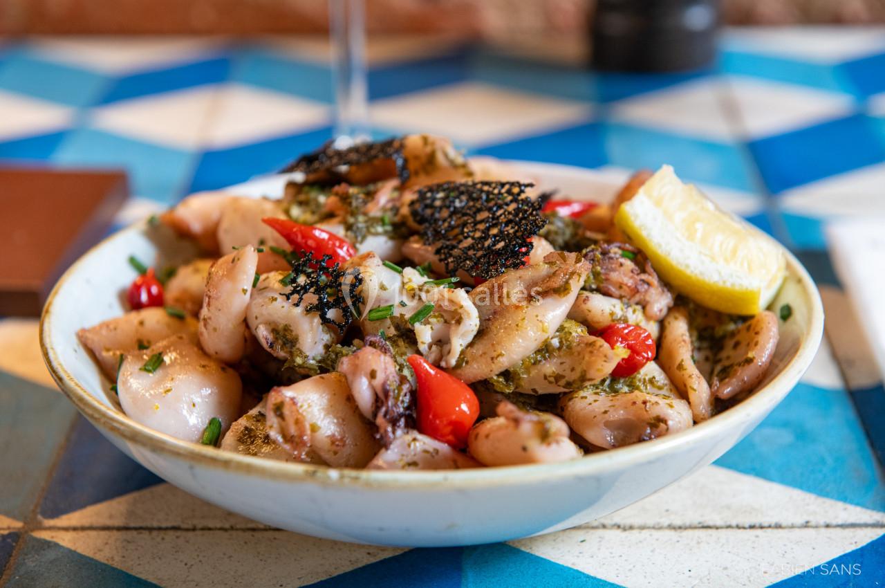 Assiette de calmars grillés garnis de tomates cerises, herbes fraîches et citron, présentée sur une table carrelée bleue.