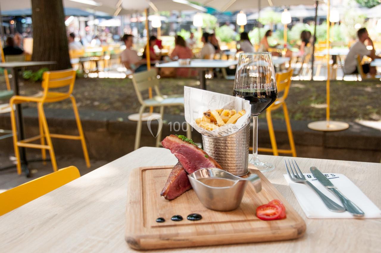 Assiette avec sandwich, frites dans un contenant métallique, sauce, verre de vin rouge, en terrasse ensoleillée.