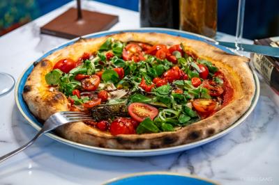 Pizza garnie de roquette, tomates cerises et légumes grillés, servie dans une assiette sur une table en marbre.