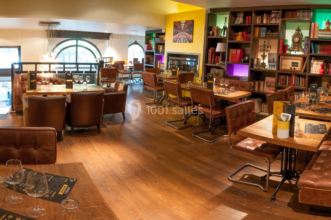 Salle de restaurant lumineuse avec tables en bois, fauteuils en cuir et étagères décorées de livres et objets.