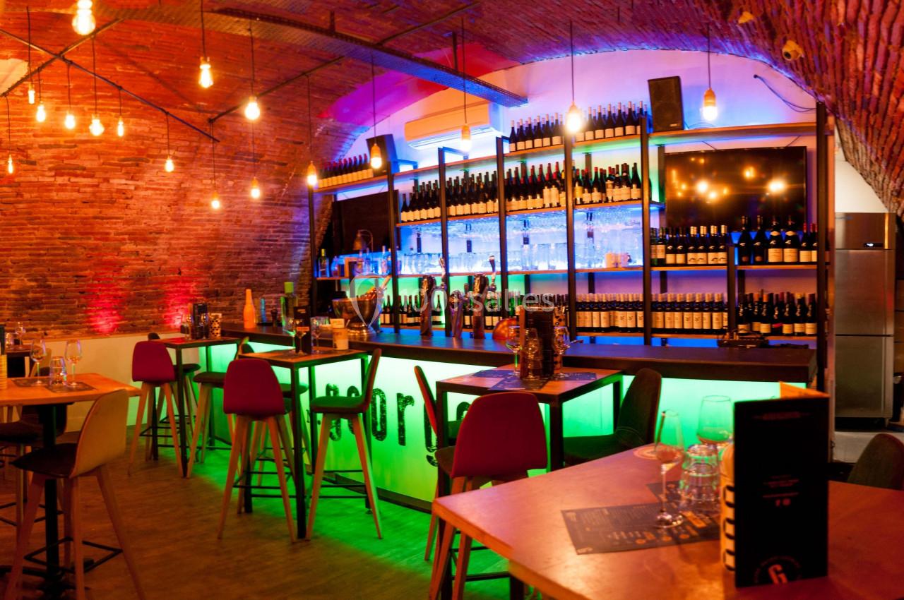 Bar moderne avec éclairage coloré, tables hautes, chaises et étagères remplies de bouteilles sous un plafond voûté en…