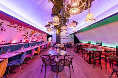 Salle de restaurant lumineuse avec banquettes bleues, chaises colorées, décor végétal et éclairage chaleureux.