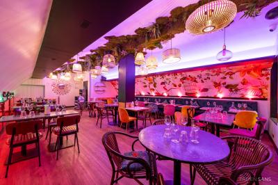 Salle de restaurant lumineuse avec banquettes bleues, chaises colorées, décor végétal et éclairage chaleureux.