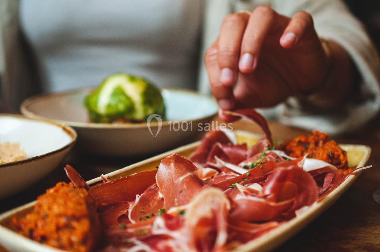 Assiette de charcuterie avec tranches de jambon cru et accompagnements, une main prenant une tranche.
