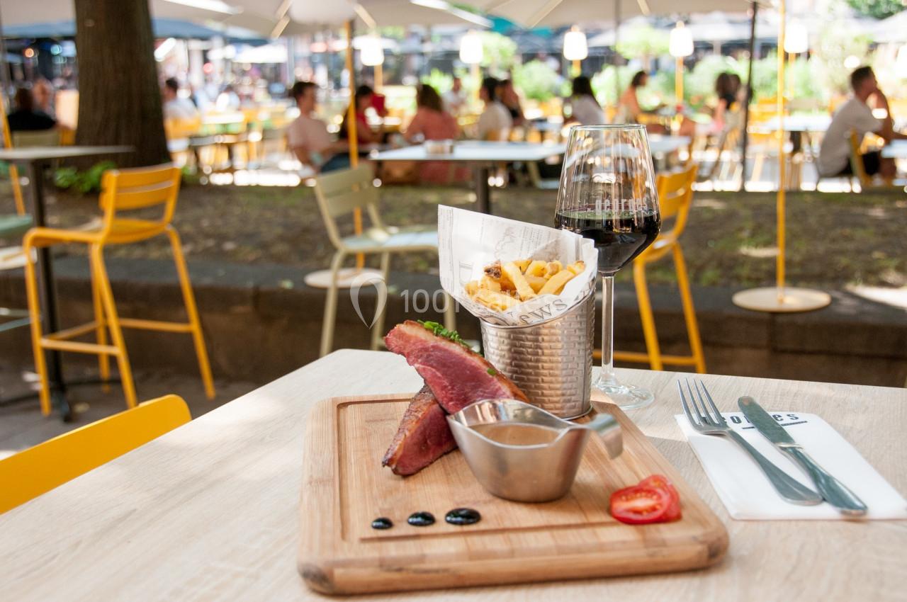Assiette avec viande grillée, frites dans un récipient métallique, verre de vin rouge, terrasse en arrière-plan.