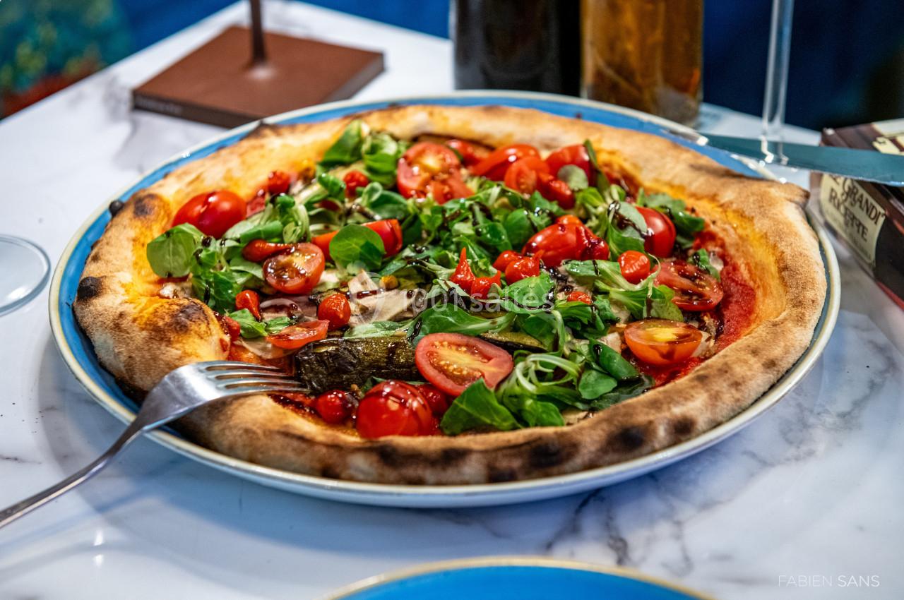 Pizza garnie de roquette, tomates cerises et légumes grillés, servie sur une assiette bleue posée sur une table en marbre.