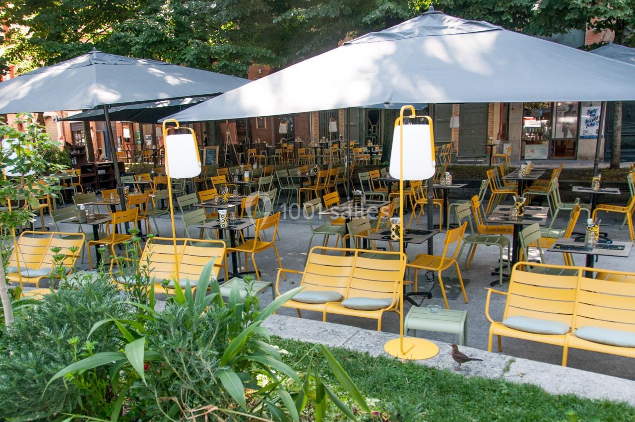 Terrasse extérieure avec des tables et des chaises jaunes sous de grands parasols gris, entourée de verdure.