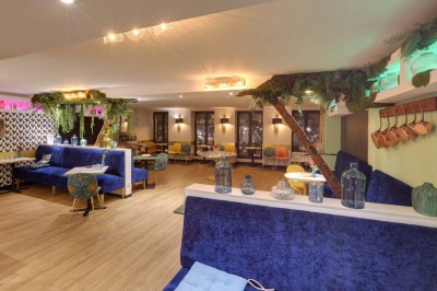 Salle de restaurant lumineuse avec banquettes bleues, chaises colorées, décor végétal et éclairage chaleureux.