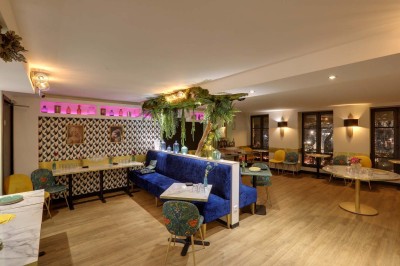 Salle de restaurant lumineuse avec banquettes bleues, chaises colorées, décor végétal et éclairage chaleureux.