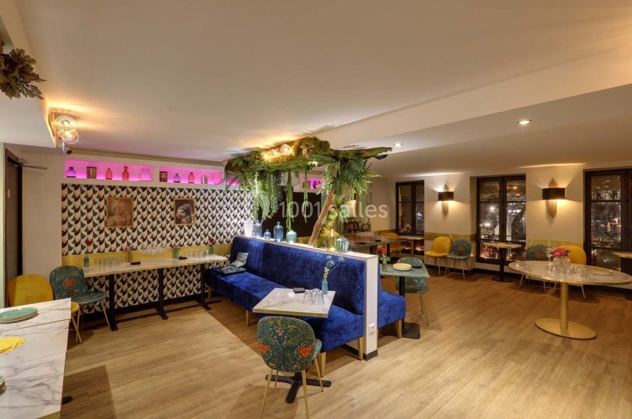 Salle de restaurant lumineuse avec banquettes bleues, chaises colorées, décor végétal et éclairage chaleureux.