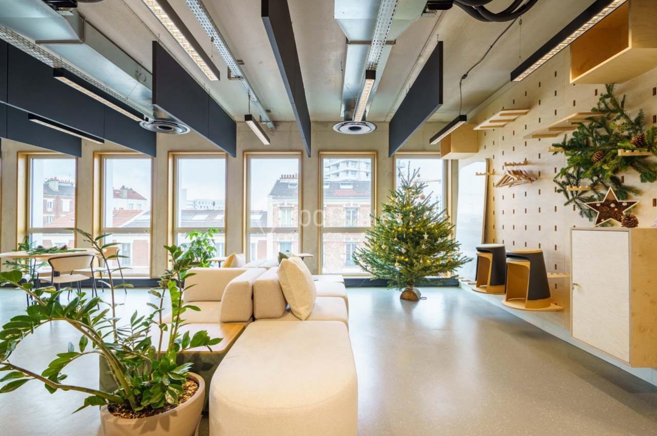 Espace intérieur lumineux avec canapé beige, plantes vertes, sapin décoré et grandes fenêtres donnant sur des bâtiments…