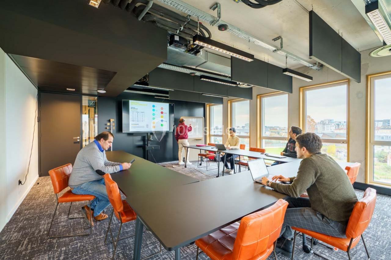 Salle de réunion moderne avec plusieurs personnes assises autour de tables, écoutant une présentation sur un écran.