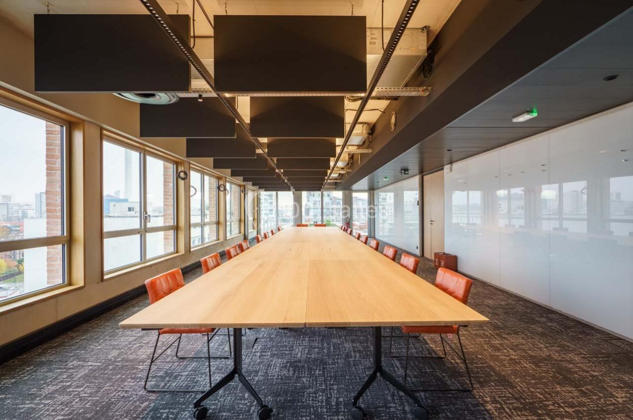 Salle de réunion lumineuse avec une grande table en bois, chaises orange et baies vitrées donnant sur la ville.