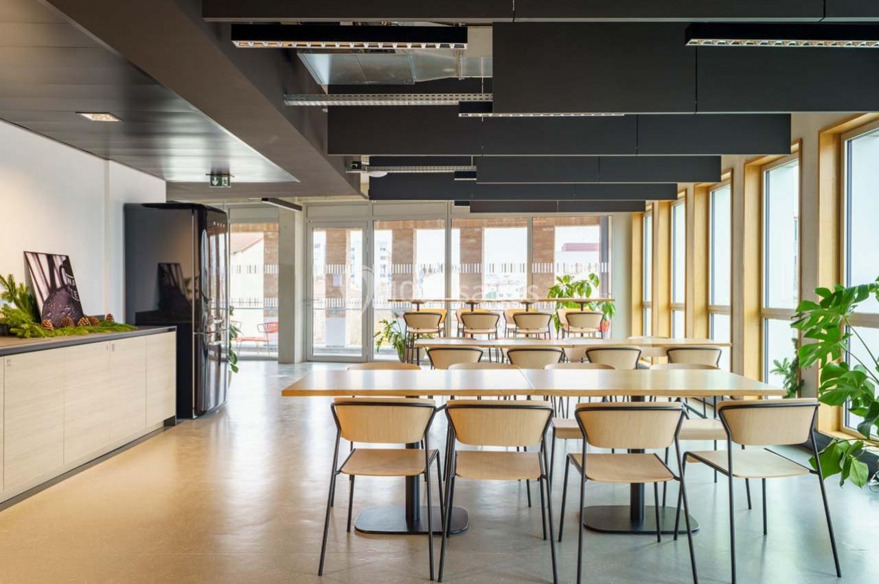 Salle lumineuse avec de grandes fenêtres, tables en bois, chaises assorties et plantes décoratives.