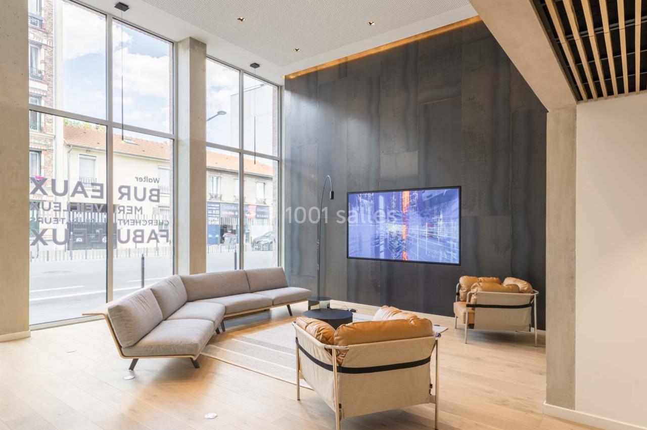 Salon moderne avec canapés et fauteuils en cuir, grande baie vitrée laissant entrer la lumière naturelle, écran mural allumé.