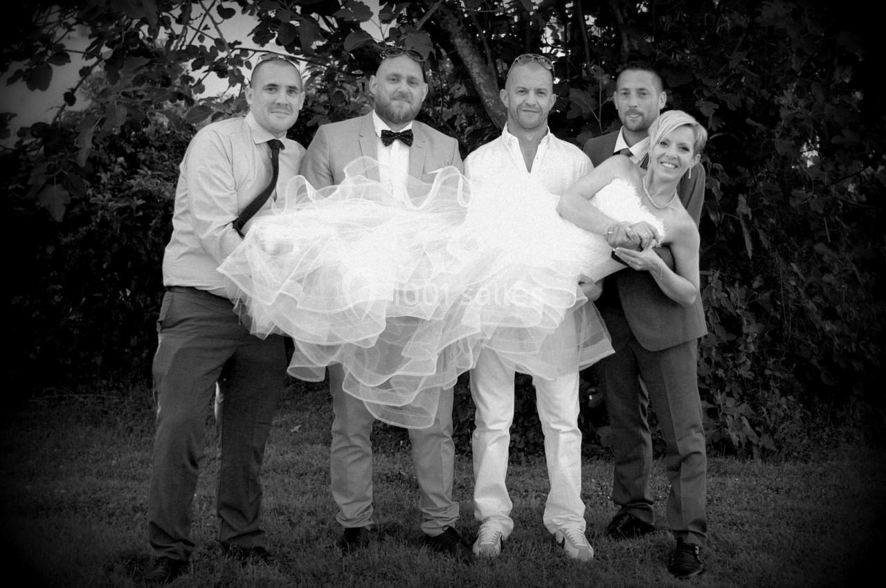 Un groupe de quatre hommes porte une femme en robe de mariée devant des arbres, tous souriants.