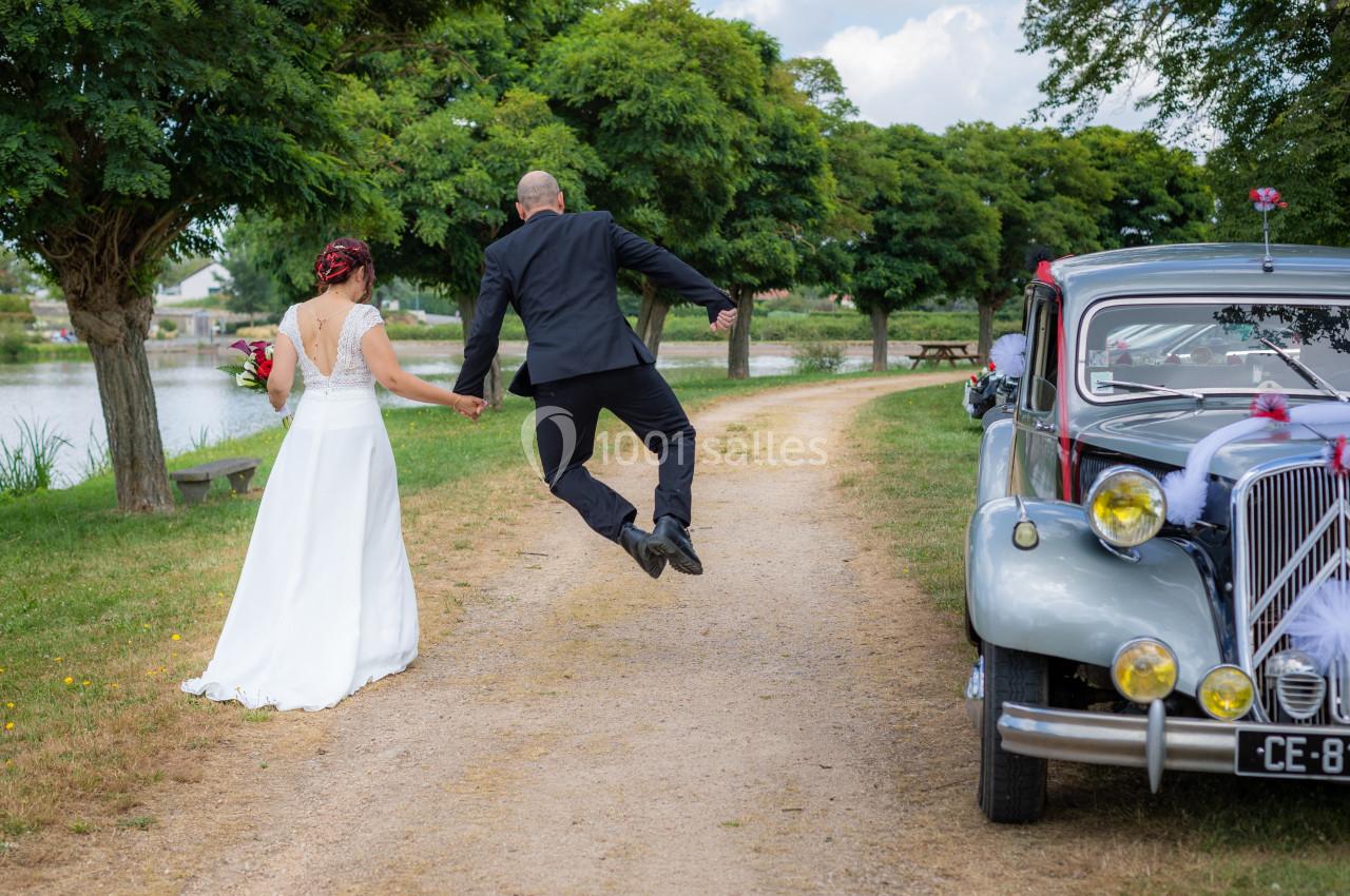 Un couple de mariés marche sur un chemin près d'une voiture ancienne, le marié saute joyeusement en l'air.