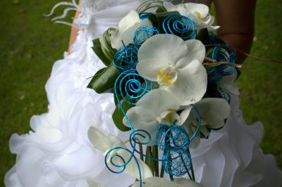 Miniature Cédric Chapelon Photographe #7 Femme en robe de mariée blanche tenant un bouquet de fleurs blanches et décorations bleues, en extérieur sur pelouse.