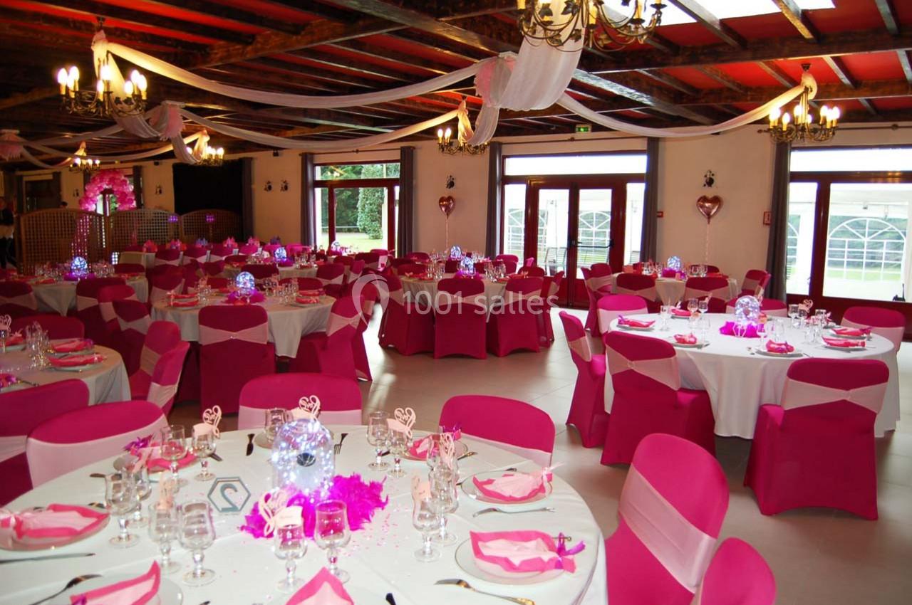 Salle de réception décorée avec des tables rondes, nappes blanches, housses de chaise roses et éclairage chaleureux.