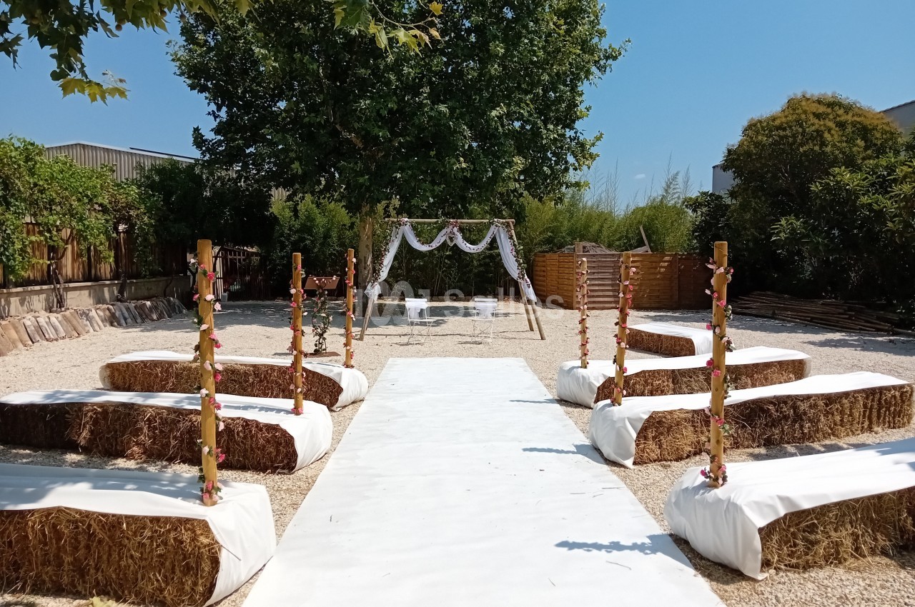 Allée centrale extérieure bordée de bottes de paille recouvertes de tissu blanc, menant à une arche décorée pour une…
