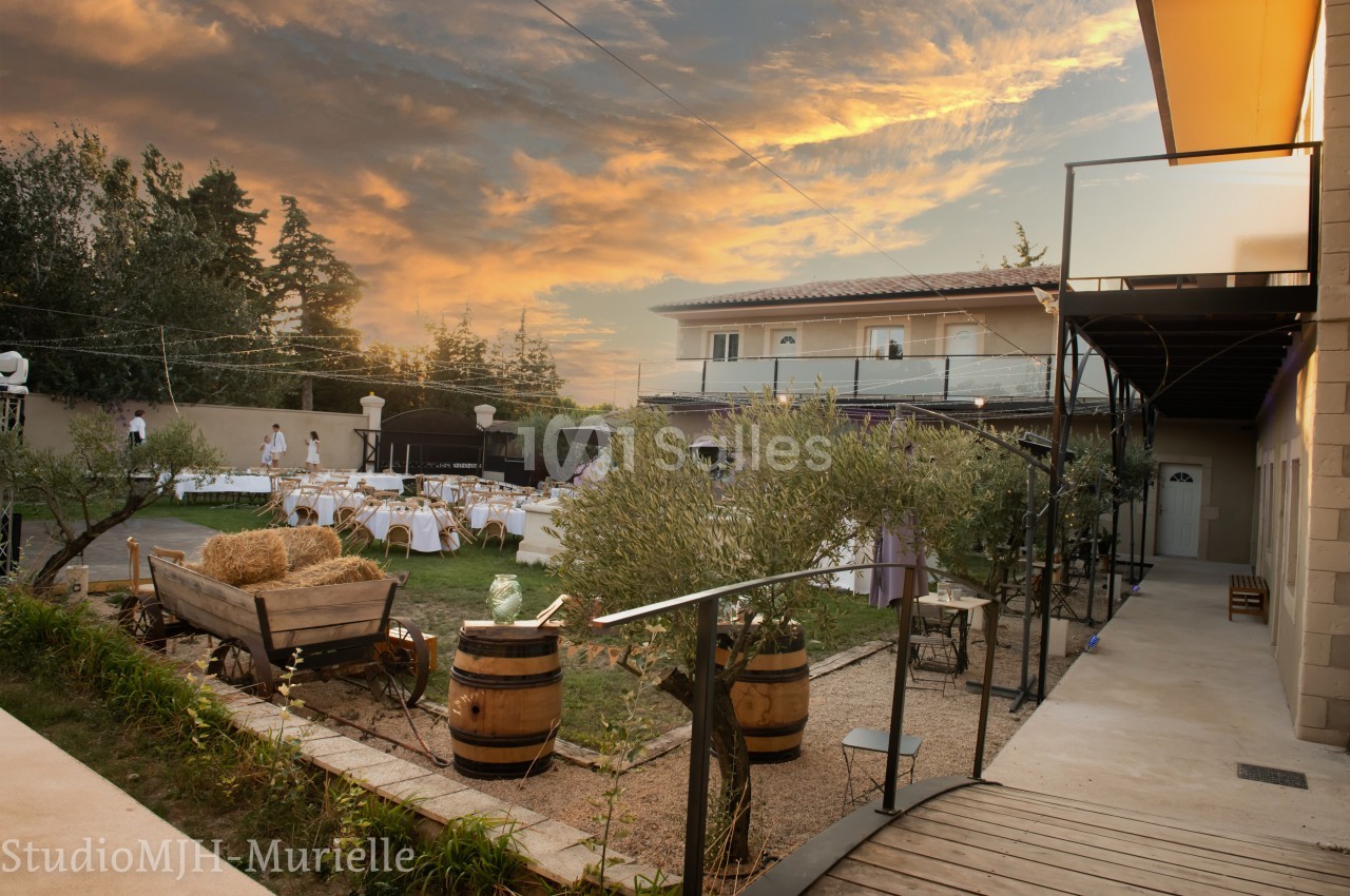 Cour extérieure aménagée pour un événement, avec tables dressées, oliviers, bottes de foin et ciel au coucher du soleil.