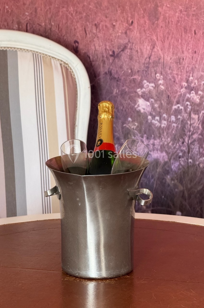 Bouteille de champagne avec deux flûtes dans un seau en métal, posés sur une table devant un fauteuil et un fond floral.