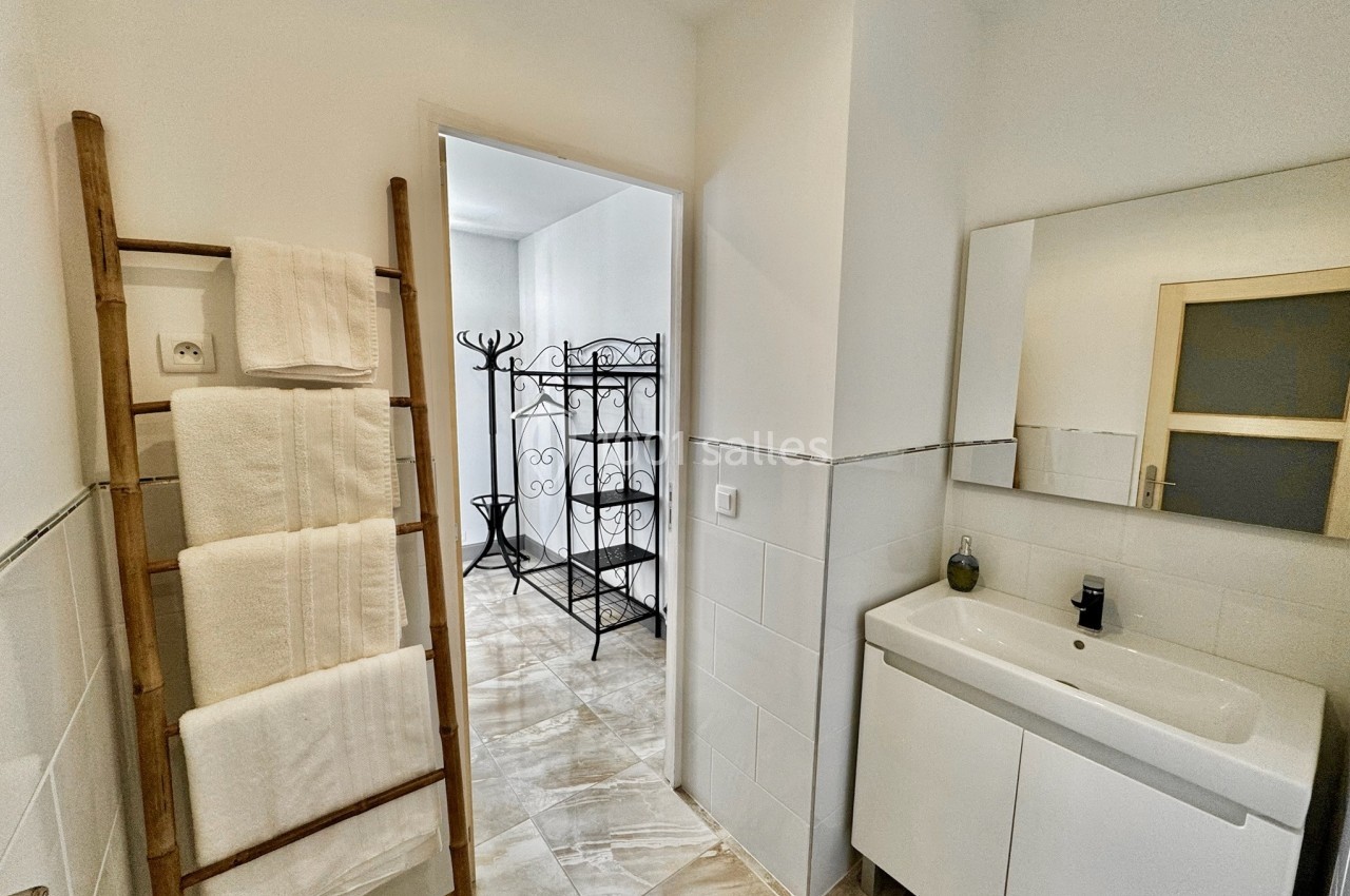 Salle de bain lumineuse avec lavabo, miroir, porte ouverte sur un espace avec porte-manteaux et échelle porte-serviettes.