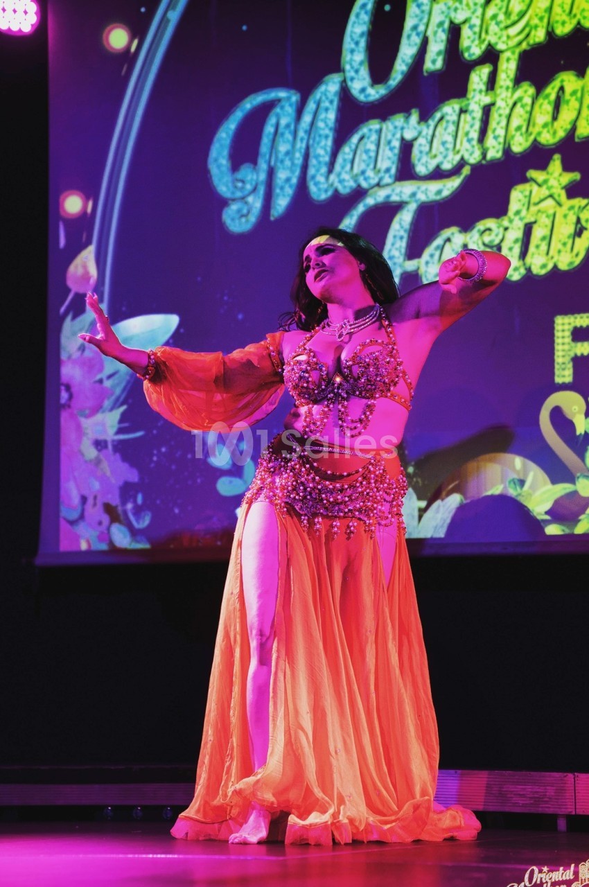 Danseuse en tenue orientale orange exécutant une performance sur une scène éclairée avec un fond coloré.