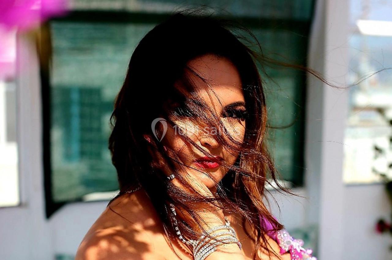 Femme aux cheveux bruns, partiellement masquée par le vent, portant des bijoux et un haut rose, en extérieur.